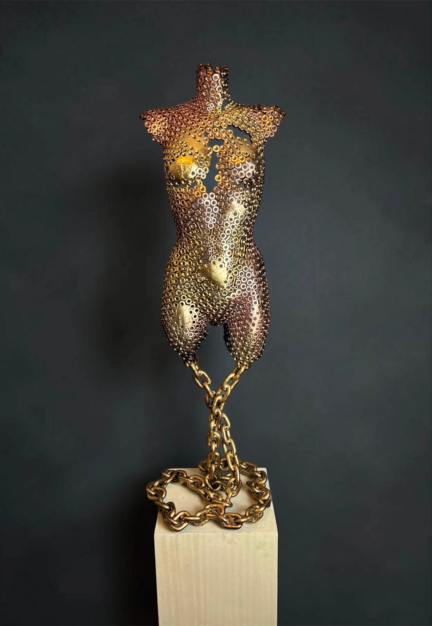 Golden Balance — goudkleurige torso van stalen moeren, hangend aan een ketting boven een betonnen sokkel